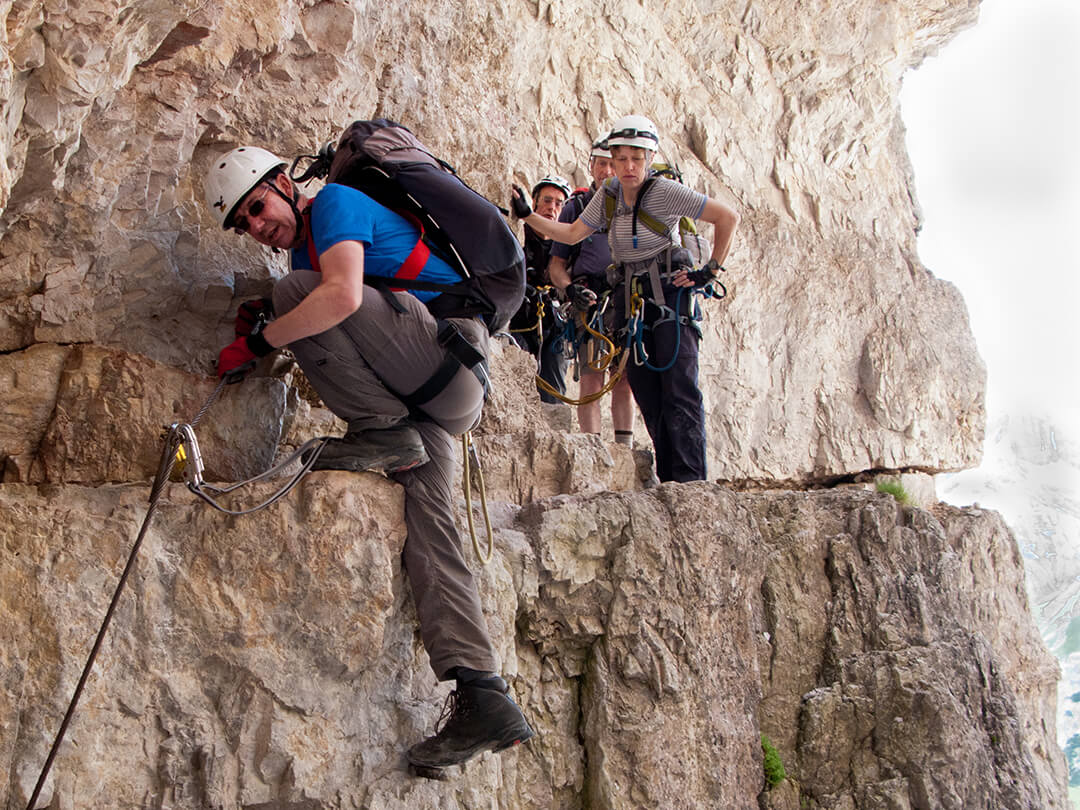 A group of people in harneses and helmets on a thin ledge on the side of a mountain clipped onto a 'Via Ferata' wire