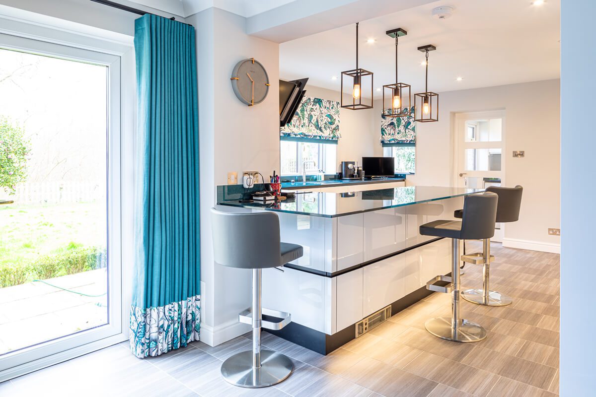 Ultra modern kitchen in white grey and blue featuring a centre island with bar stools. Highlighted are the blue full length curtains and roman blinds